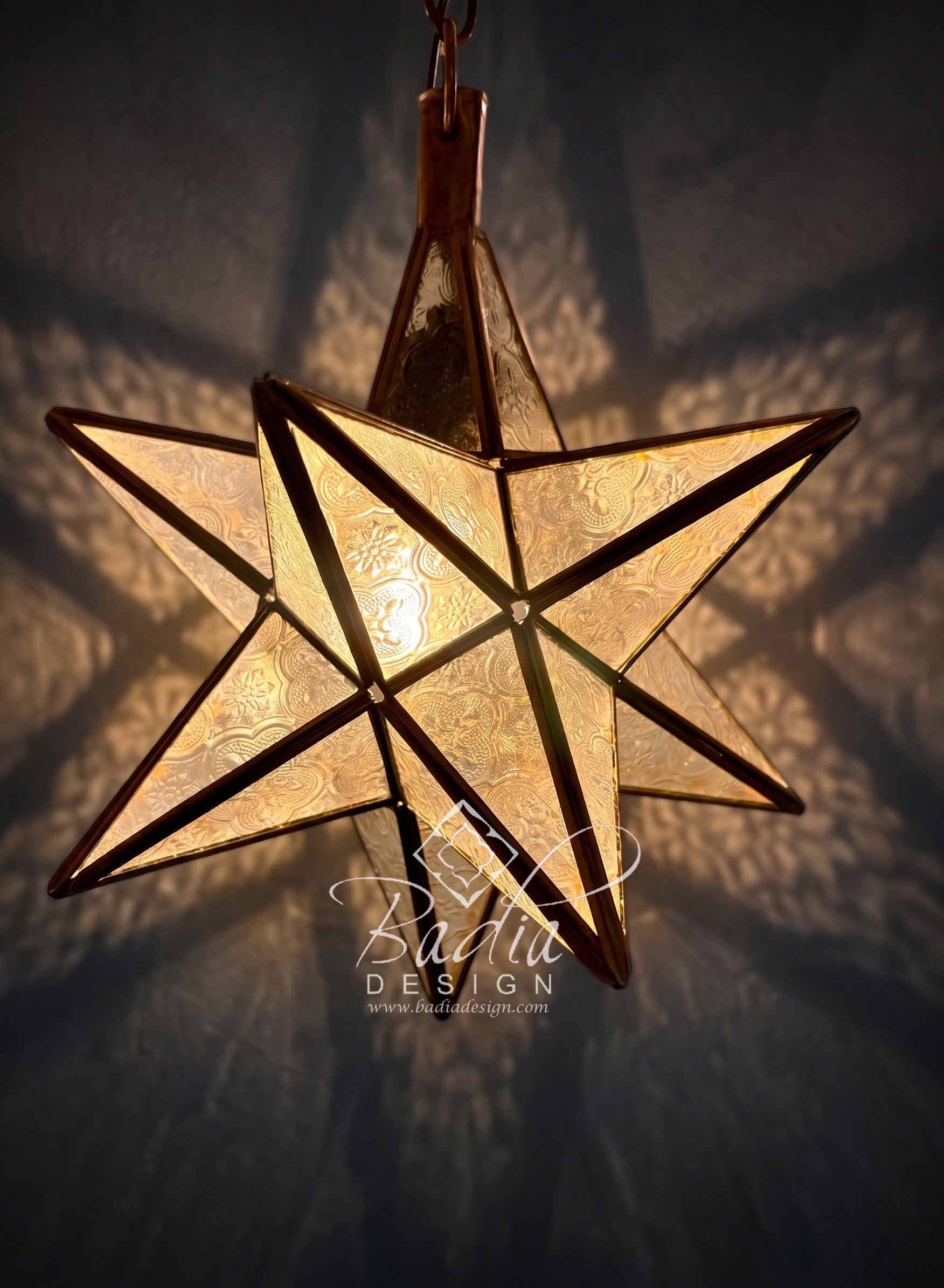 A hanging five-pointed star pendant light with textured frosted glass panels and dark metal framing, glowing warmly against a dark wall with shadow patterns.