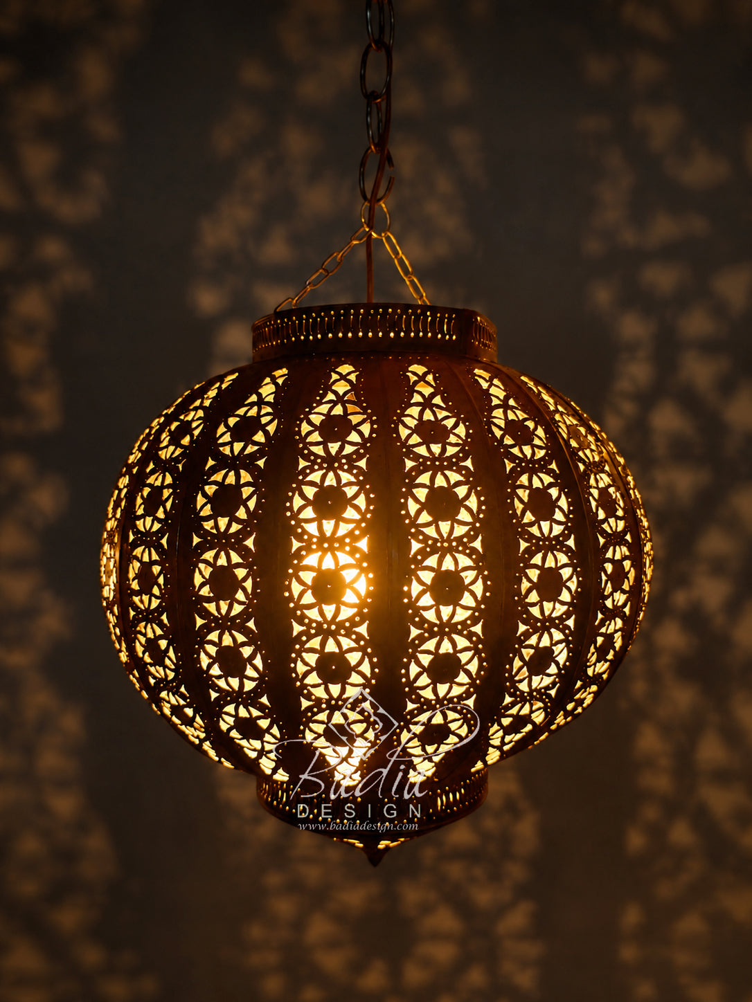 A spherical Moroccan-style hanging pendant lamp with intricate laser-cut geometric patterns, glowing warmly from an internal light bulb. The lamp is suspended by a chain against a dark background illuminated with star-like shadow patterns.