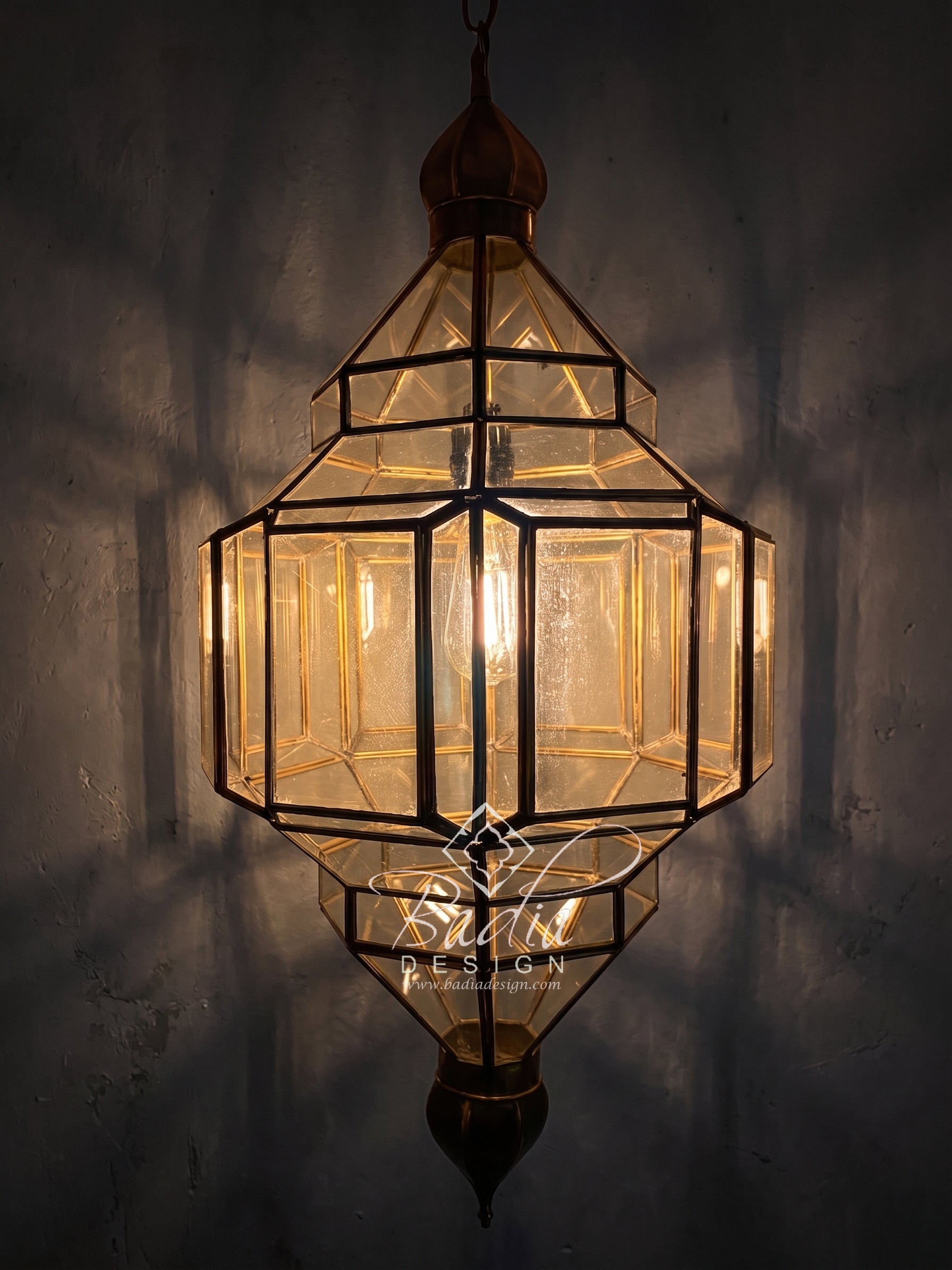 A geometric hanging lantern with clear glass panels and dark metal framing, illuminated from within against a dark textured wall. A watermark reading &
