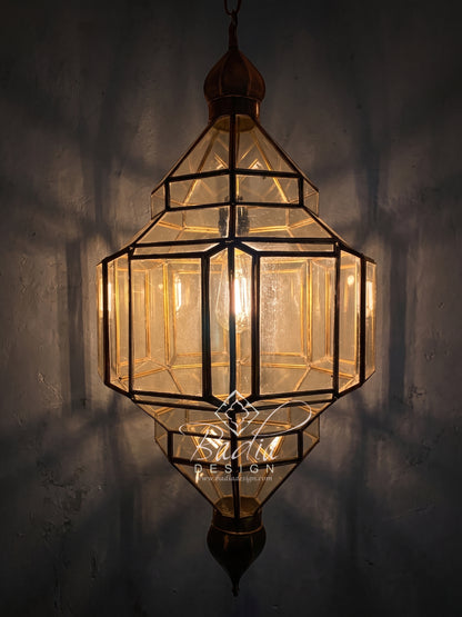 A geometric hanging lantern with clear glass panels and dark metal framing, illuminated from within against a dark textured wall. A watermark reading &