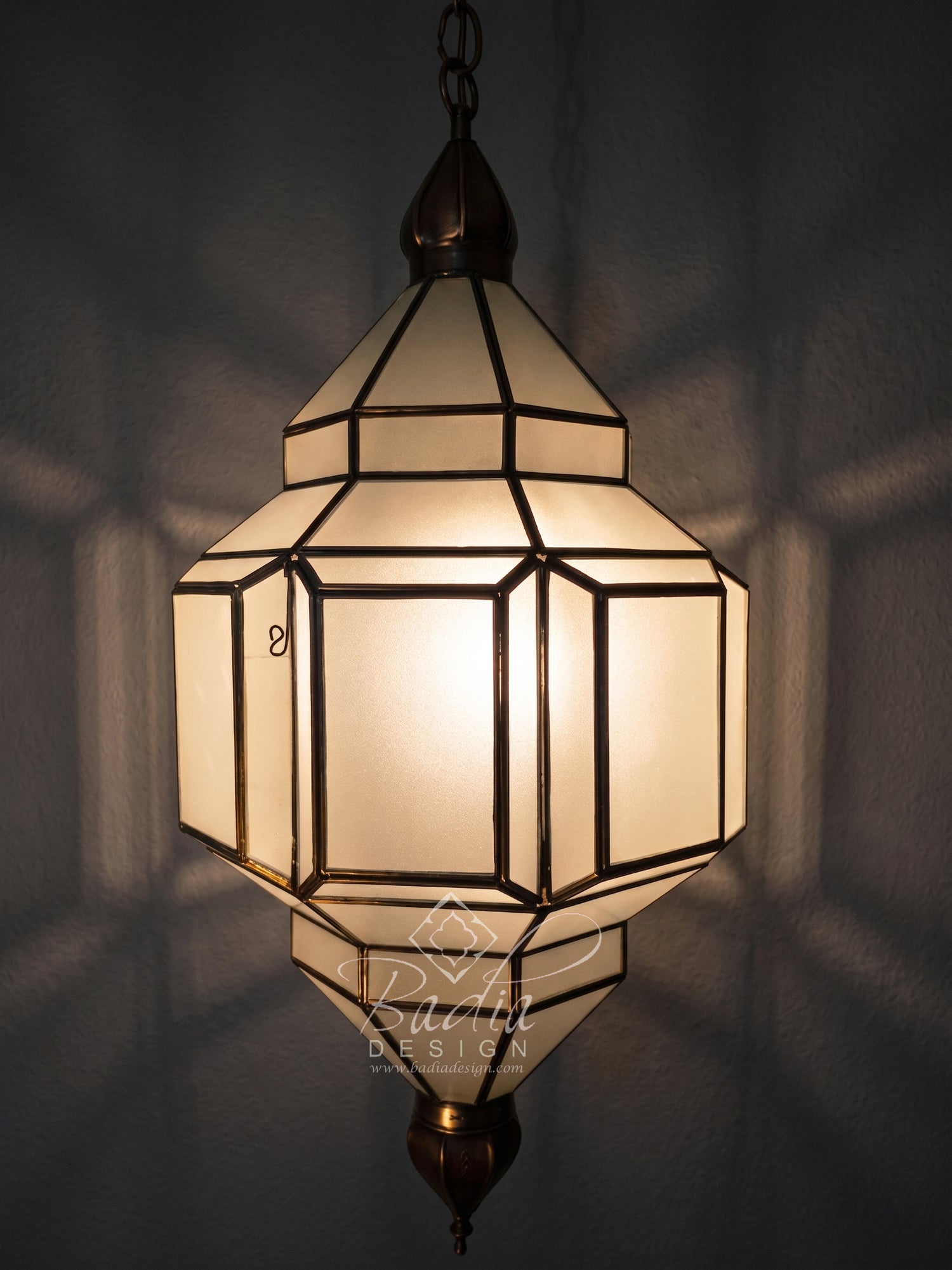 A geometric hanging lantern-style pendant light with frosted white panels and dark bronze metal framing, shown illuminated against a dark wall with soft shadow patterns.