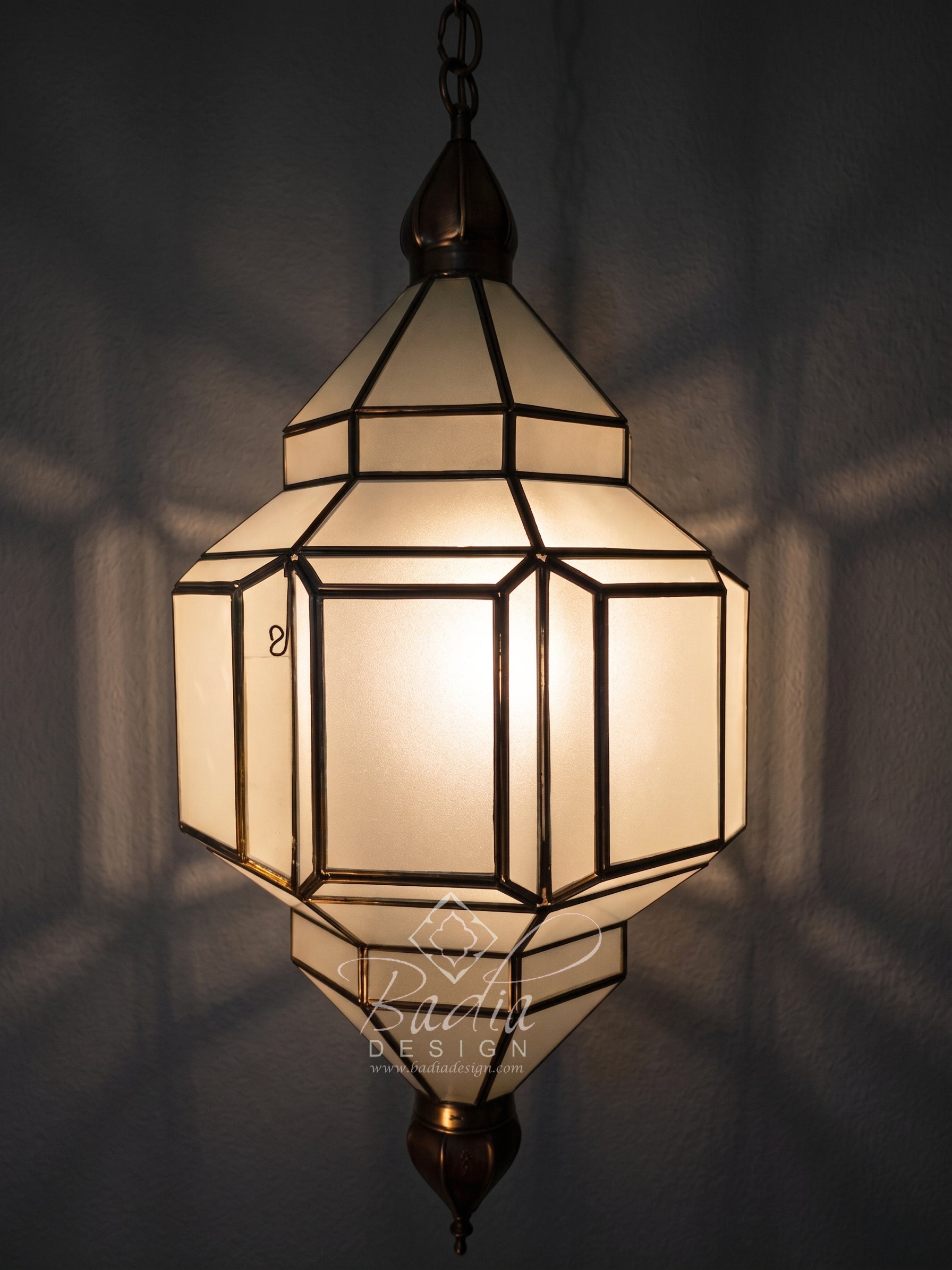 A geometric hanging lantern-style pendant light with frosted white panels and dark bronze metal framing, shown illuminated against a dark wall with soft shadow patterns.