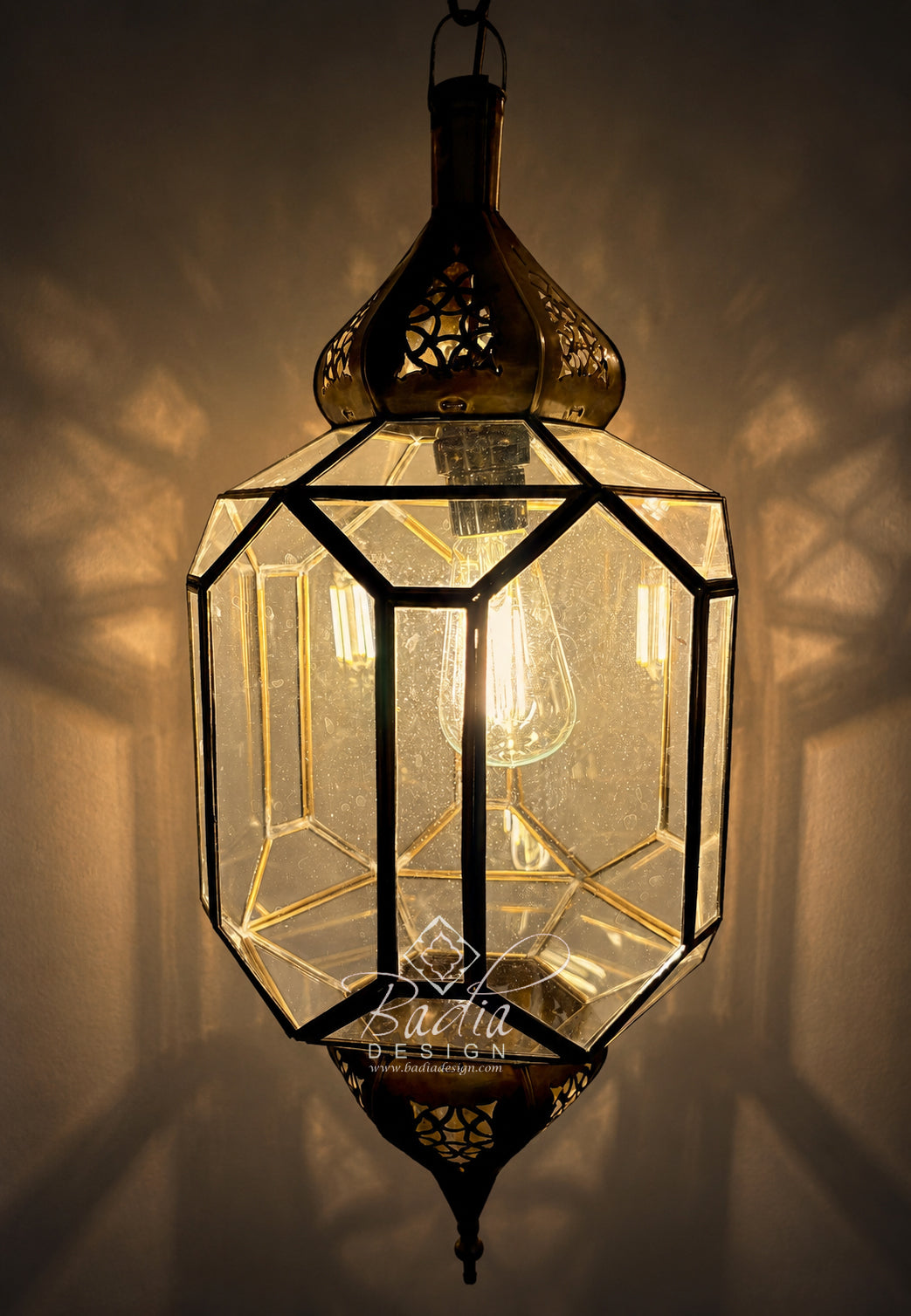 A geometric brass hanging lantern with clear glass panels and a visible bulb inside, casting warm light against a wall. A watermark reading &