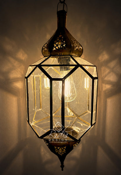 A geometric brass hanging lantern with clear glass panels and a visible bulb inside, casting warm light against a wall. A watermark reading &