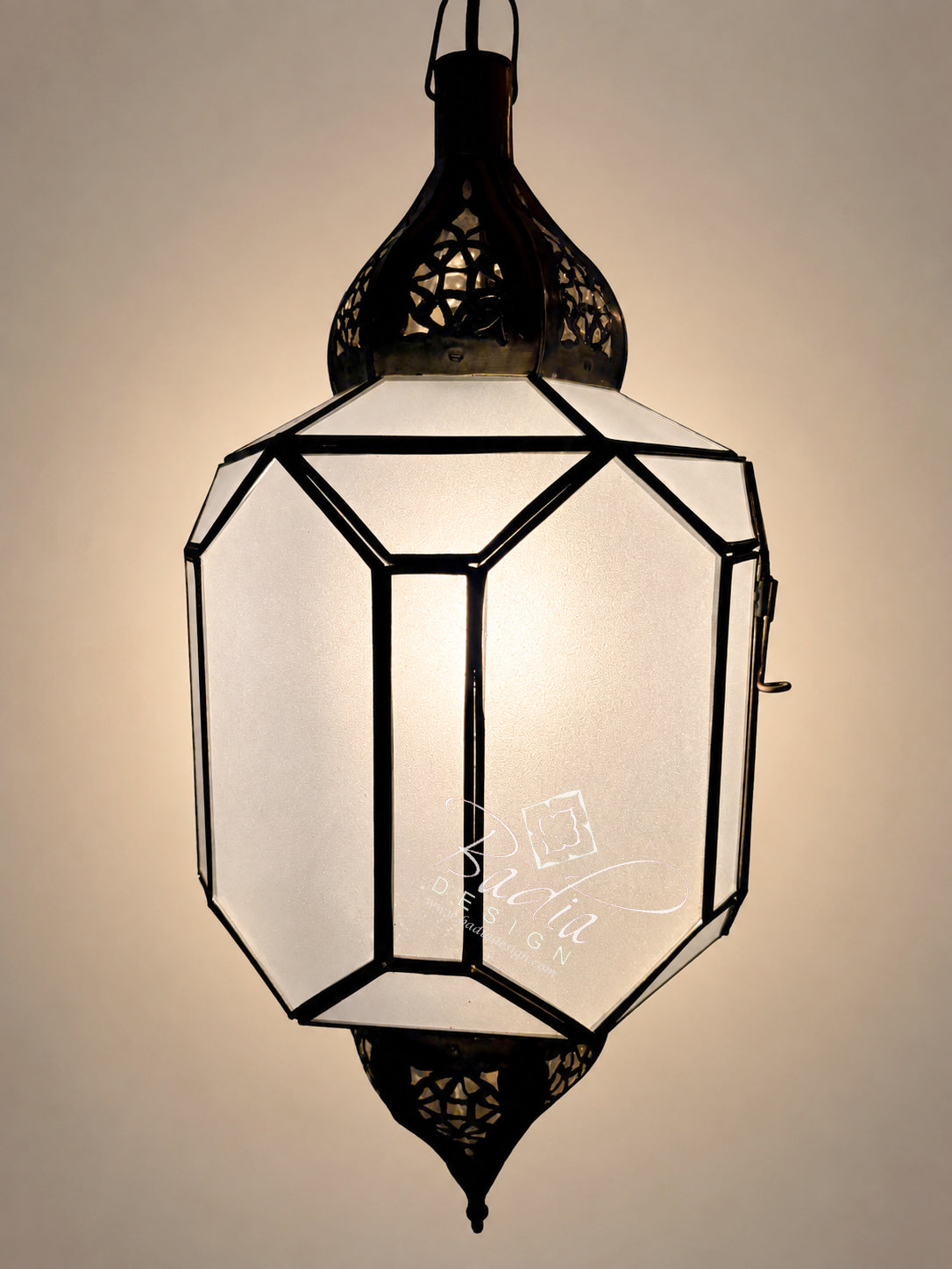 A hanging Moroccan-style lantern with a black metal frame and white frosted glass panels, glowing with warm light against a beige background.