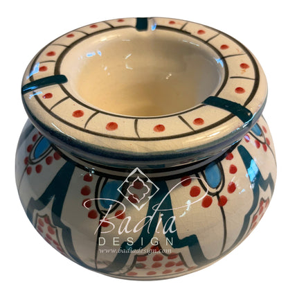 A small round ceramic ashtray with a wide rim, decorated with a cream glaze, blue geometric patterns, and red dots, shown on a white background with a &