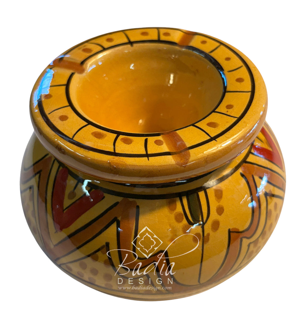 A small round ceramic pot or candle holder with a glossy yellow-orange glaze, decorated with black geometric lines and red and brown patterns. The pot has a wide rim with circular dotted details and is shown on a white background with a &