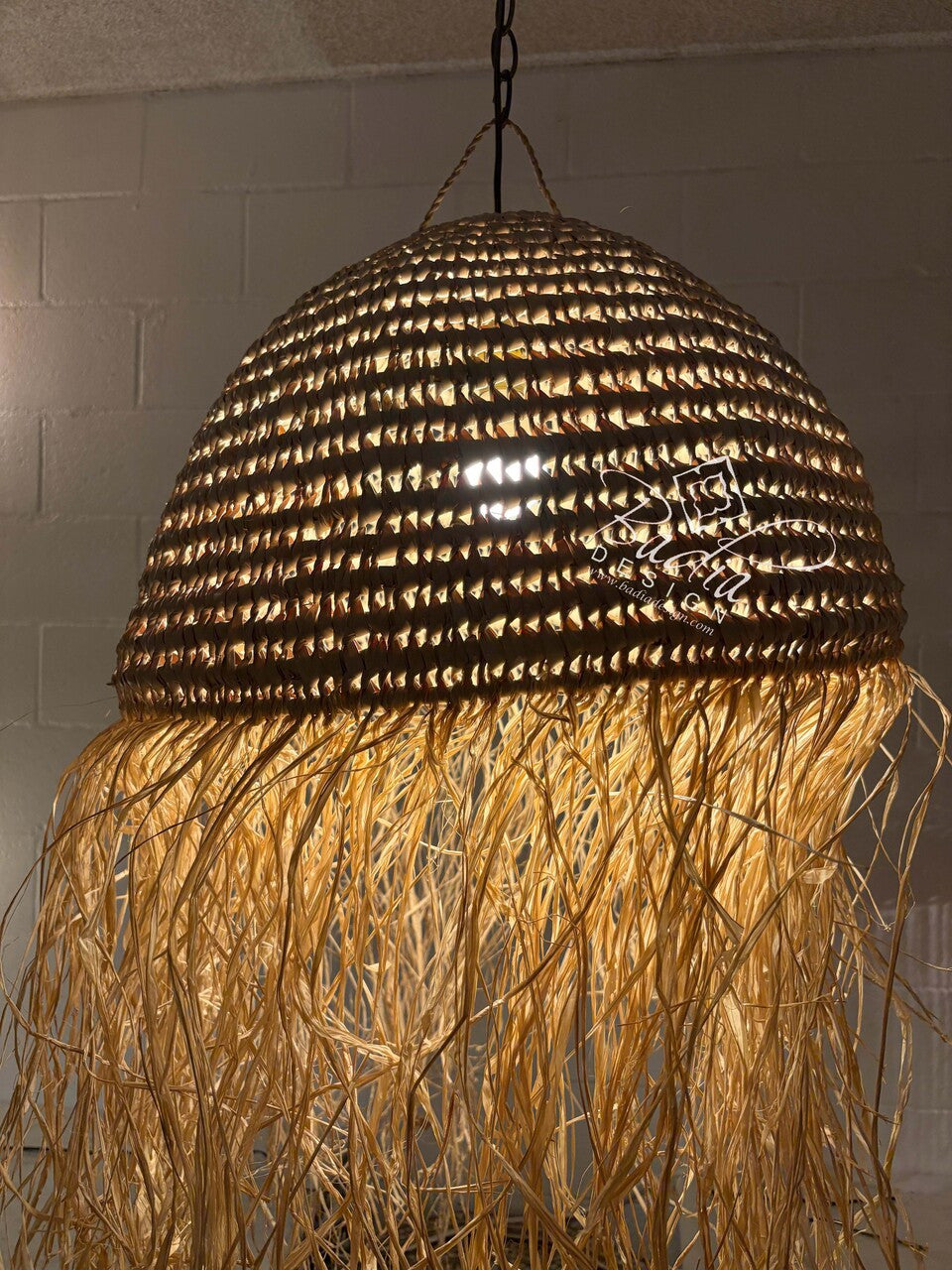Woven pendant light with natural fiber fringe against a tiled wall.