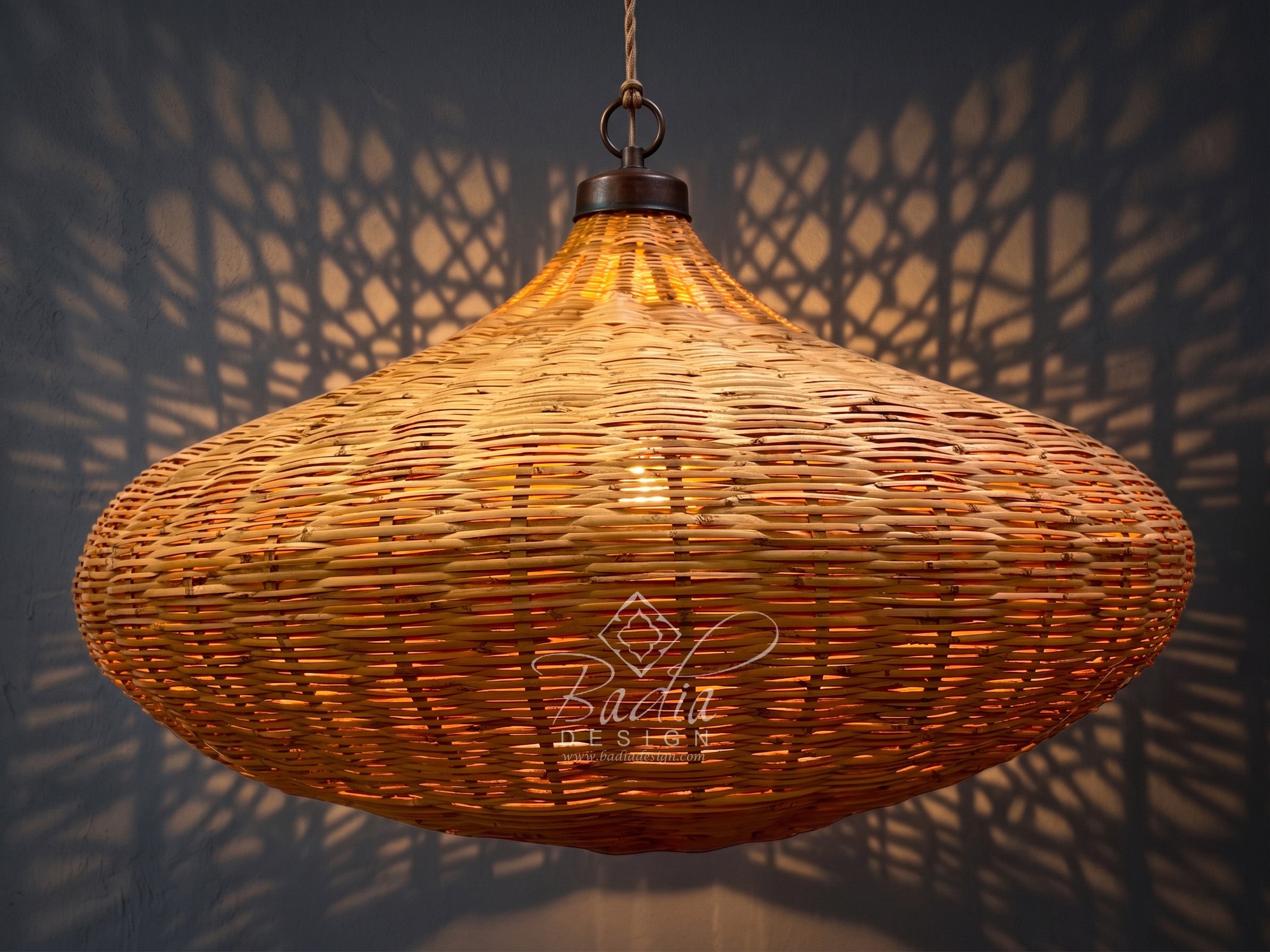 A large woven rattan pendant light in a teardrop shape, glowing with warm light and casting geometric shadows on a dark wall. A &