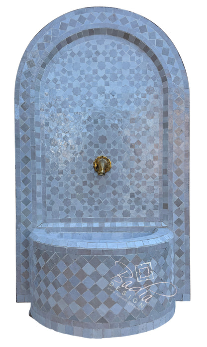 An arched mosaic fountain wall with a curved basin, featuring light blue and gray tiles with a central gold lion-head spout, shown on a plain white background.