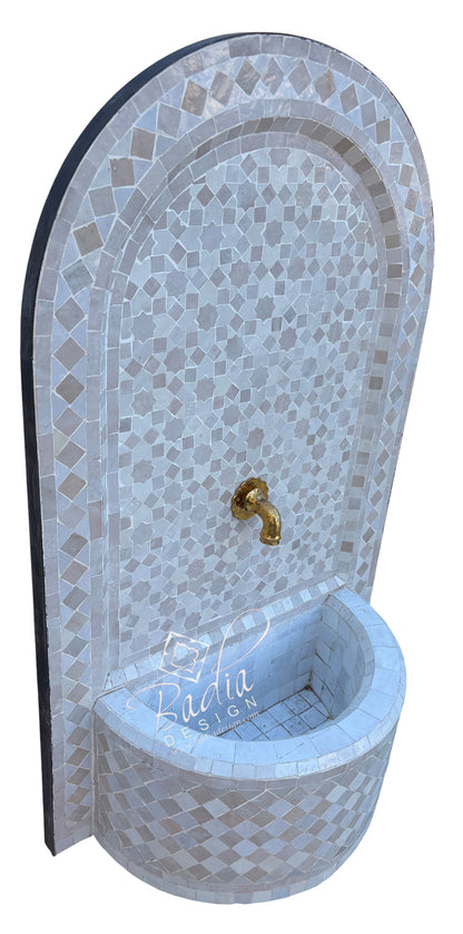 An arched mosaic fountain wall basin with a curved back panel, featuring light gray and white tiles, a small gold faucet, and a white tiled basin at the bottom.