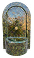 An arched mosaic fountain wall with a matching cylindrical basin, covered in reflective green, blue, and gold tiles. A small lion-head spout is centered near the top.
