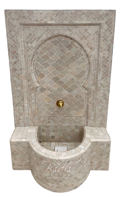 A Moorish-style mosaic wall fountain with a curved basin, beige and gray scalloped tile pattern, and a small gold faucet, shown against a plain white background.
