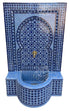 A Moroccan-style mosaic wall fountain with a curved basin, featuring intricate blue and white tile patterns, a central gold faucet, and a white background.