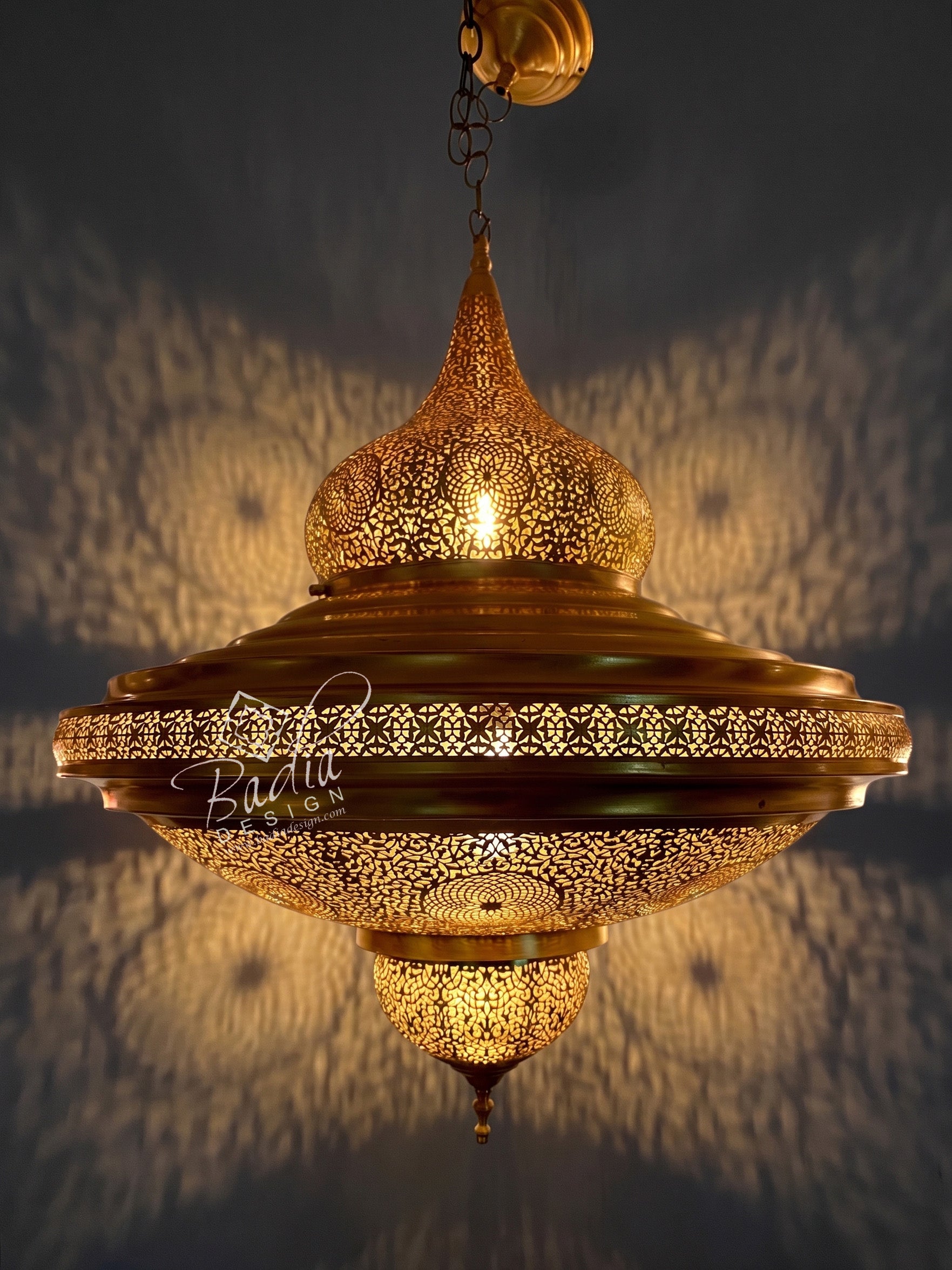 An ornate brass hanging pendant lamp with intricate laser-cut geometric patterns, glowing with warm light against a dark wall that casts soft shadow patterns.