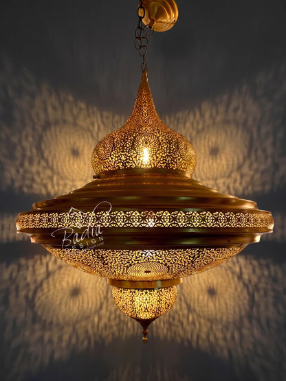 An ornate brass hanging pendant lamp with intricate laser-cut geometric patterns, glowing with warm light against a dark wall that casts soft shadow patterns.