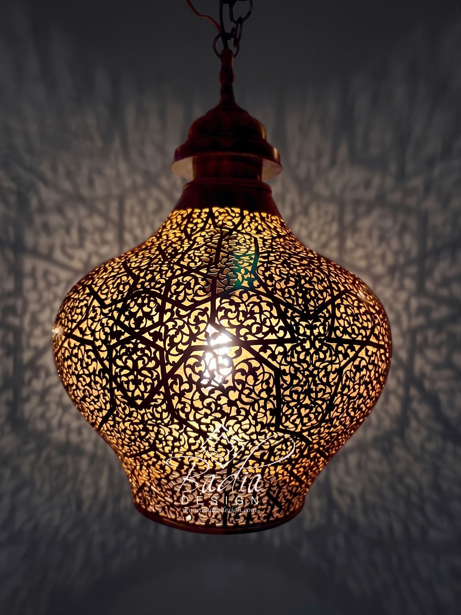 A hanging Moroccan-style metal pendant lamp with an intricate cutout pattern, glowing warmly from within against a dark wall that casts shadow patterns.