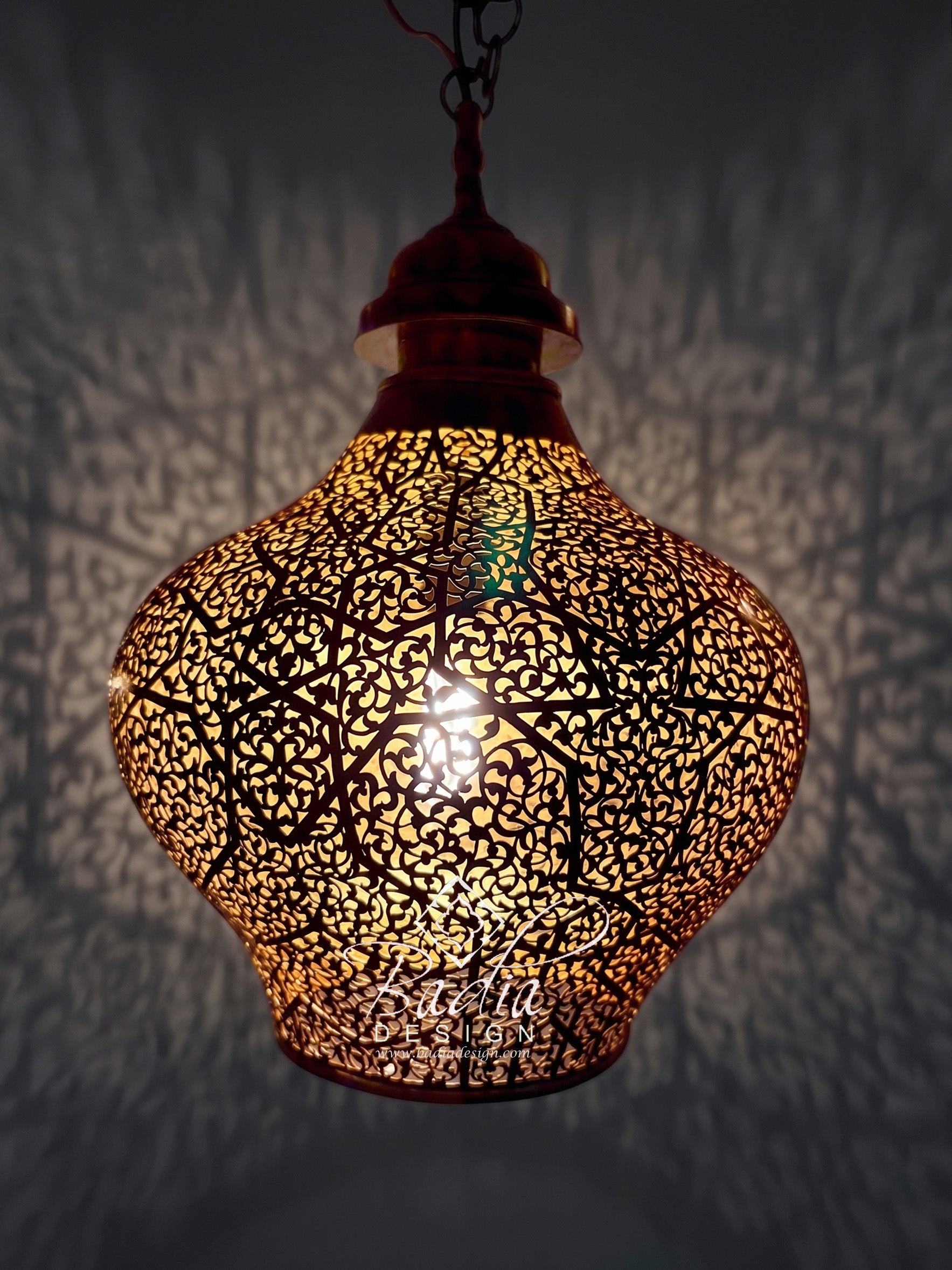A hanging Moroccan-style metal pendant lamp with an intricate cutout pattern, glowing warmly from within against a dark wall that casts shadow patterns.