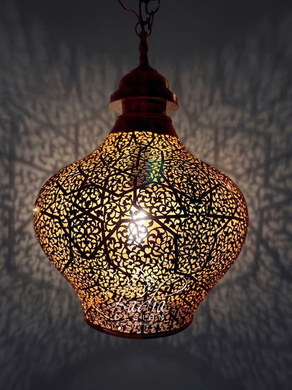 A hanging Moroccan-style metal pendant lamp with an intricate cutout pattern, glowing warmly from within against a dark wall that casts shadow patterns.