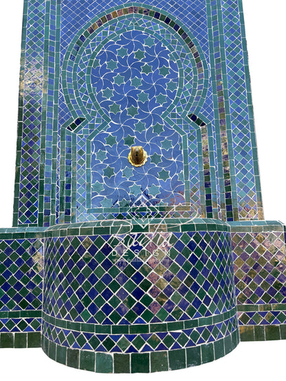 A close-up of a mosaic tile wall or fountain panel featuring geometric and floral patterns in shades of blue, teal, green, and white, with a small gold-toned circular detail near the top center.
