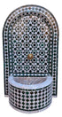 A Moroccan-style mosaic wall fountain with an arched back panel and a curved basin, decorated in green, white, black, and gray tile patterns with a central brass spout.