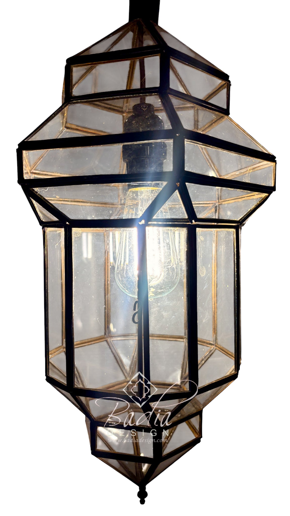 A geometric lantern-style pendant light with a black metal frame and clear glass panels, shown against a plain white background. A warm light glows from within, and a watermark reading &