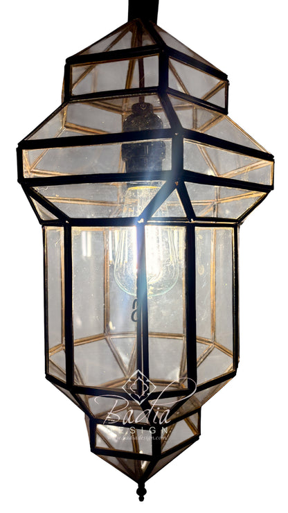 A geometric lantern-style pendant light with a black metal frame and clear glass panels, shown against a plain white background. A warm light glows from within, and a watermark reading &