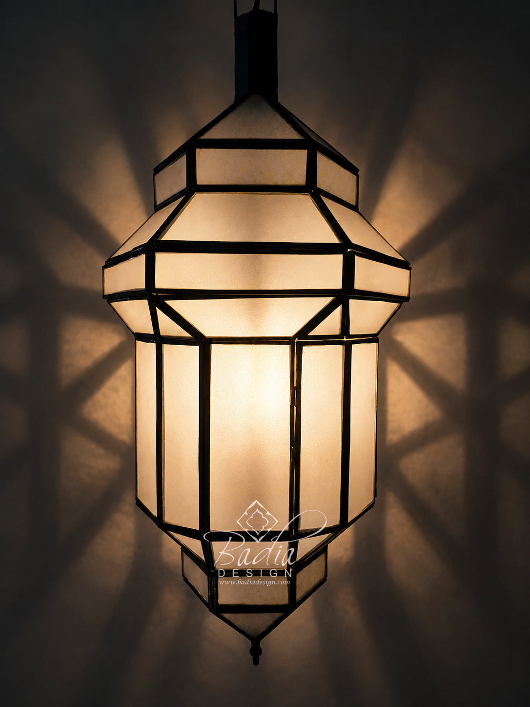 A geometric hanging lantern with warm glowing frosted glass panels and dark metal framing, shown against a dark wall with dramatic shadow patterns cast outward. A visible watermark reads &