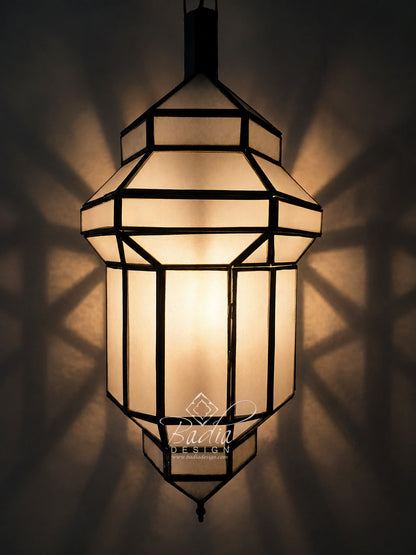 A geometric hanging lantern with warm glowing frosted glass panels and dark metal framing, shown against a dark wall with dramatic shadow patterns cast outward. A visible watermark reads &