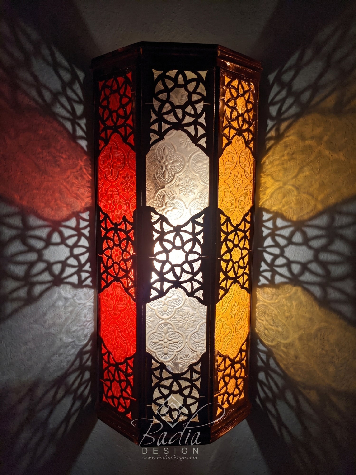 A tall hexagonal Moroccan-style wall lamp with intricate black metal cutout patterns and red, white, and yellow stained glass panels, casting dramatic shadows on a textured wall.