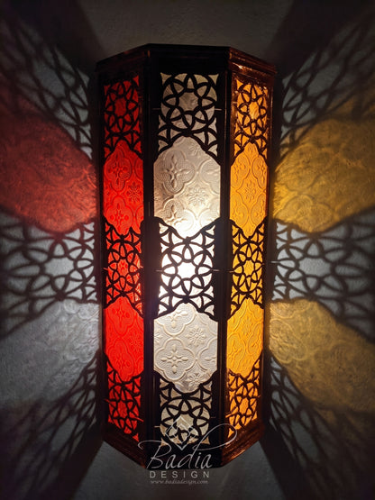 A tall hexagonal Moroccan-style wall lamp with intricate black metal cutout patterns and red, white, and yellow stained glass panels, casting dramatic shadows on a textured wall.
