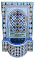 A Moroccan-style mosaic wall fountain with a curved tiled basin, decorative geometric patterns, and a central arch motif. The design features blue, green, white, brown, and gold tiles with a brass faucet at the top.