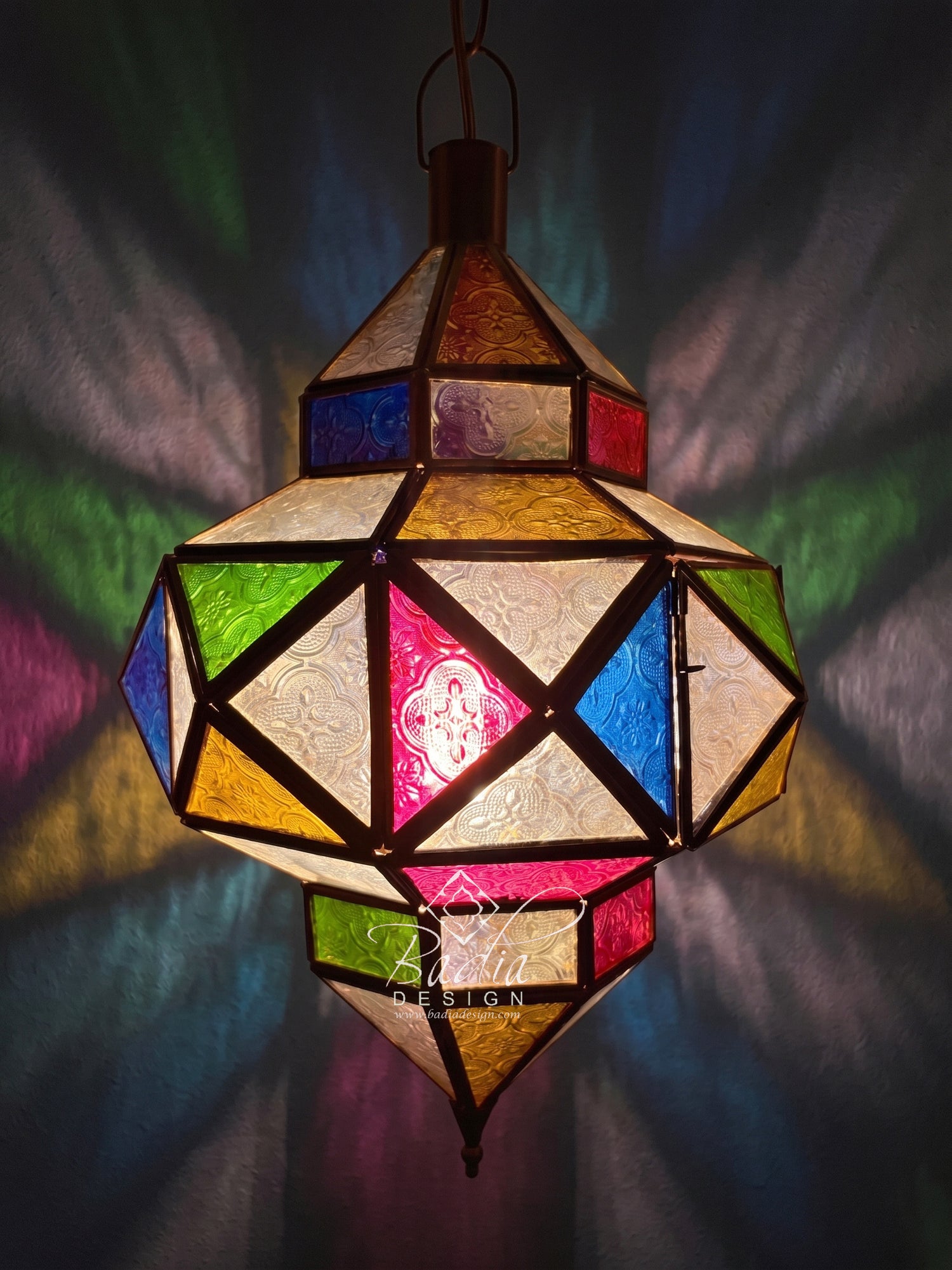 A hanging geometric stained-glass lantern with multicolored panels in blue, green, yellow, pink, and cream, casting colorful light patterns on a dark background. The brand name &