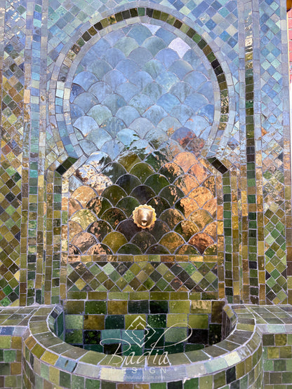 Closeup of A Moroccan-style mosaic tile fountain wall with an arched opening, green and blue scalloped tiles, a lion head detail, and a curved basin below.