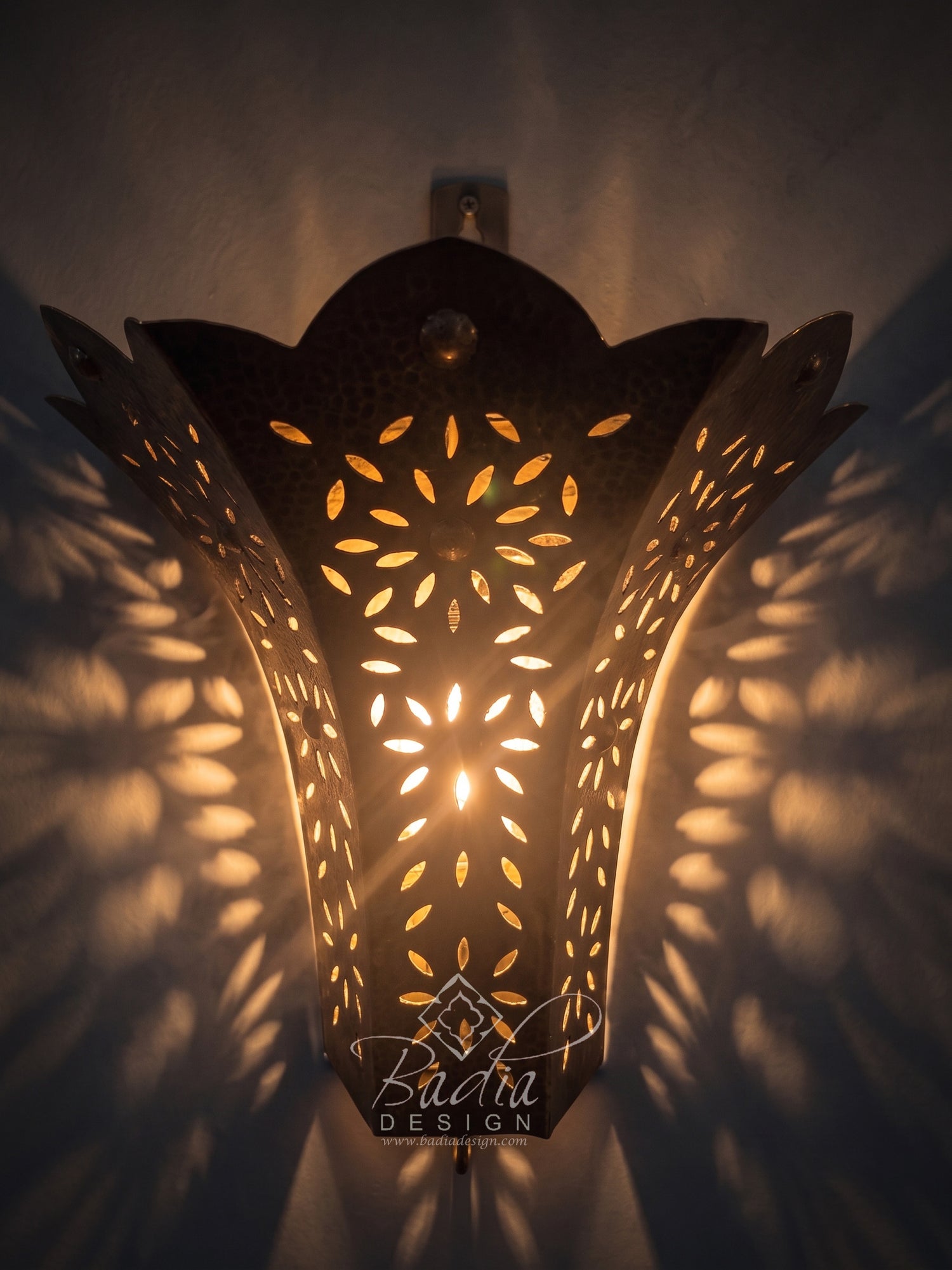 A decorative metal wall sconce with intricate cutout patterns, casting warm golden light and shadow patterns on a textured wall. The brand name &