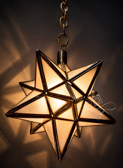 A hanging star-shaped pendant light with a warm glowing illuminated glass center and dark metal frame, suspended from a chain against a dark background with cast shadows.