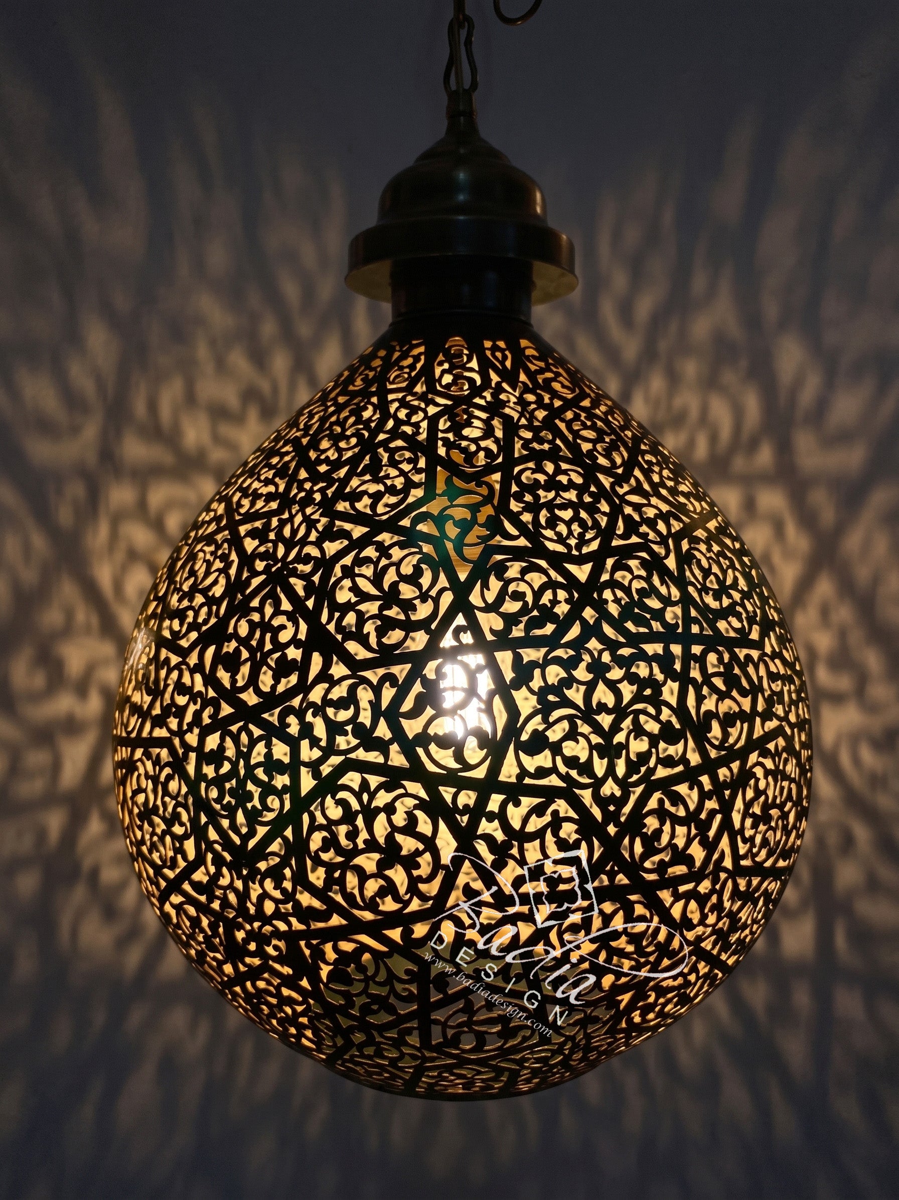 A spherical black metal Moroccan-style pendant lamp with intricate cutout patterns, glowing with warm light against a dark wall that casts leaf-like shadows.