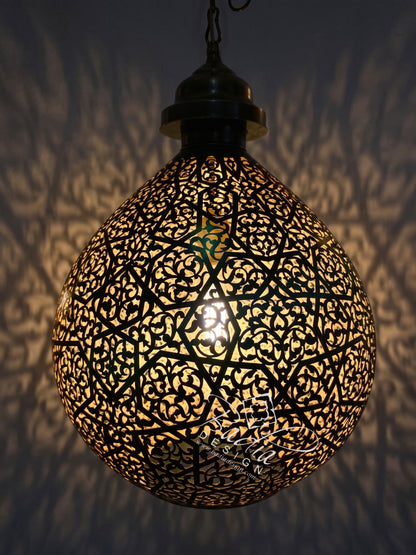 A spherical black metal Moroccan-style pendant lamp with intricate cutout patterns, glowing with warm light against a dark wall that casts leaf-like shadows.