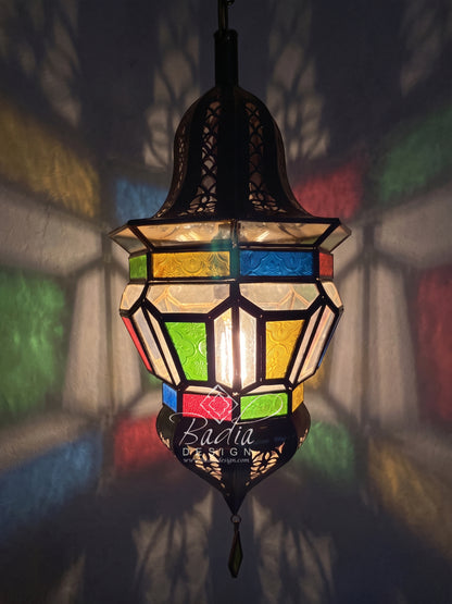 A hanging Moroccan-style stained glass lantern with a black metal frame and multiple colorful glass panels, glowing with warm light against a dark wall with colorful shadows.