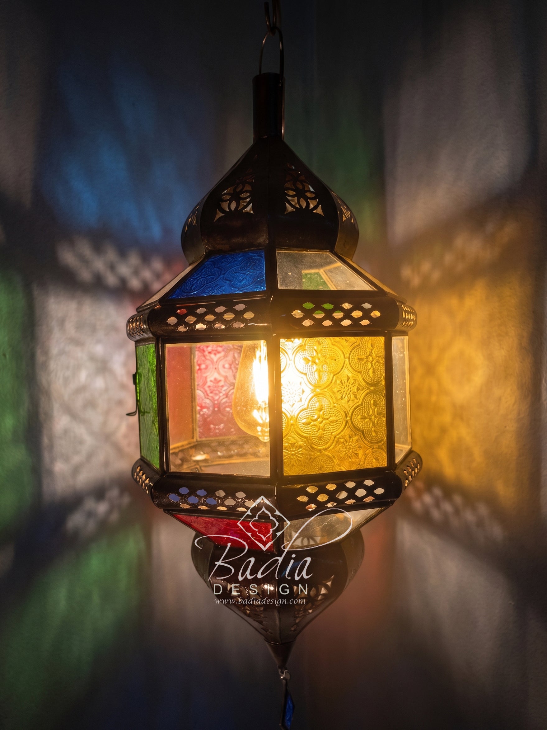 A hanging Moroccan-style lantern with a black metal frame and colorful stained glass panels, glowing with a warm light against a dark background with colored shadows.
