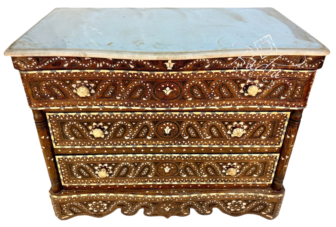 An ornate three-drawer wooden chest of drawers with intricate mother-of-pearl inlay work in brown, gold, and cream, topped with a light blue-gray marble slab, shown on a white background.