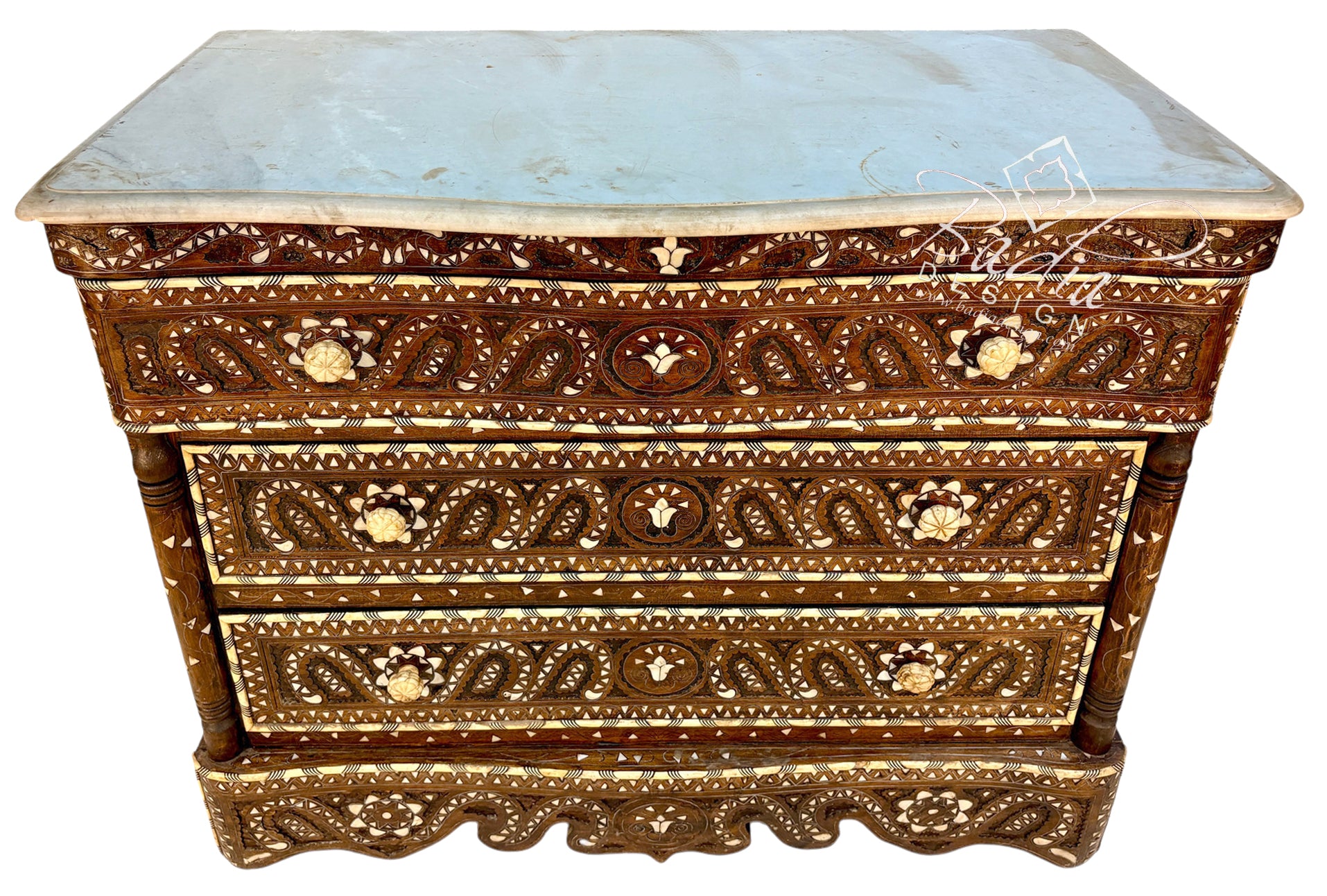 An ornate three-drawer wooden chest of drawers with intricate mother-of-pearl inlay work in brown, gold, and cream, topped with a light blue-gray marble slab, shown on a white background.