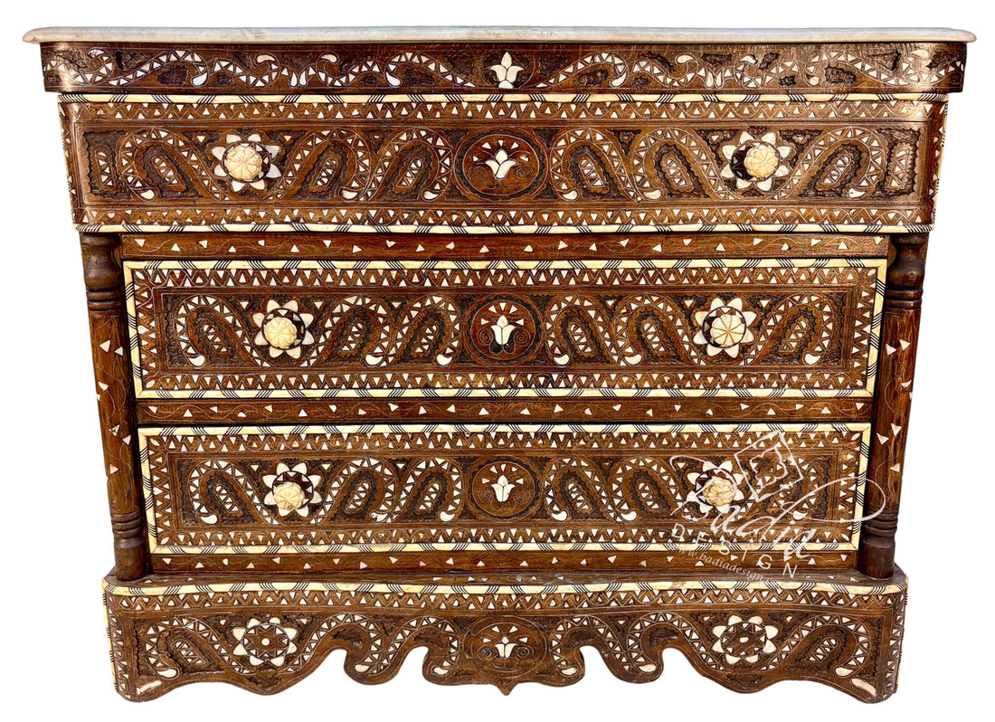 An ornate carved wooden chest of drawers with intricate mother-of-pearl inlays, featuring three drawers and a carved apron, shown against a plain white background.