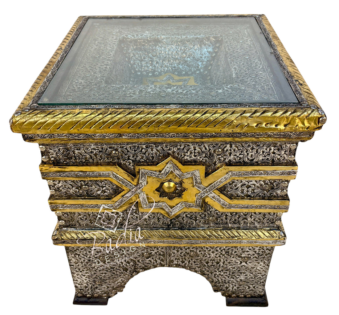 An ornate square side table with a glass top, featuring intricate silver filigree and gold accents, set against a plain white background.