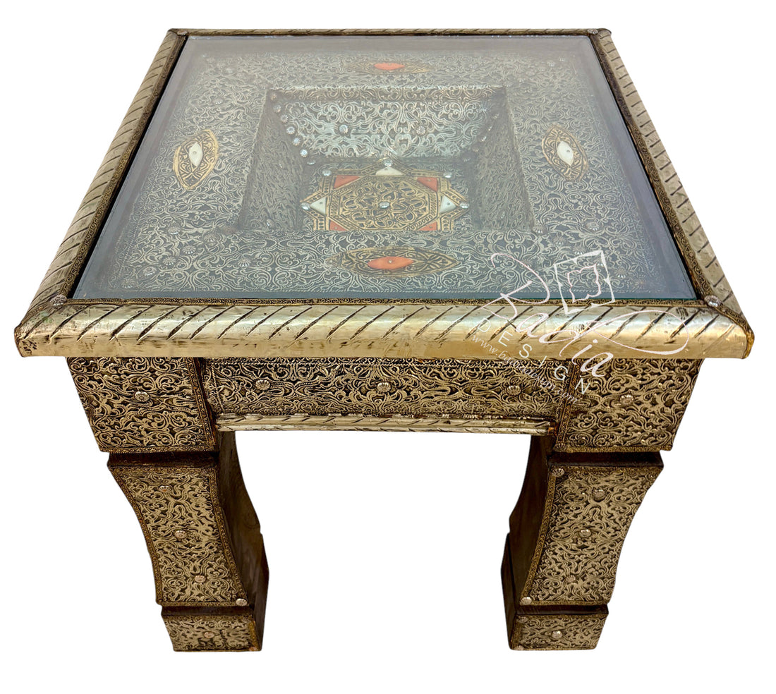 An ornate square side table with a carved brass or bronze metal frame, featuring a clear glass top and intricate floral inlay patterns in gold, silver, and red on the tabletop.