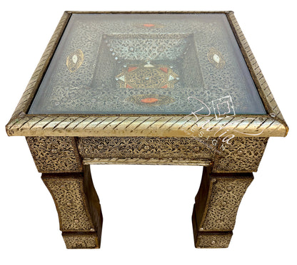 An ornate square side table with a carved brass or bronze metal frame, featuring a clear glass top and intricate floral inlay patterns in gold, silver, and red on the tabletop.