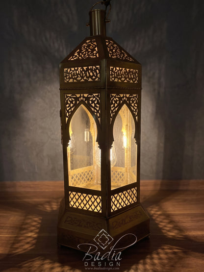 An ornate brass Moroccan-style lantern with intricate cutout patterns, illuminated from within to cast warm light on a wooden surface against a dark textured wall.