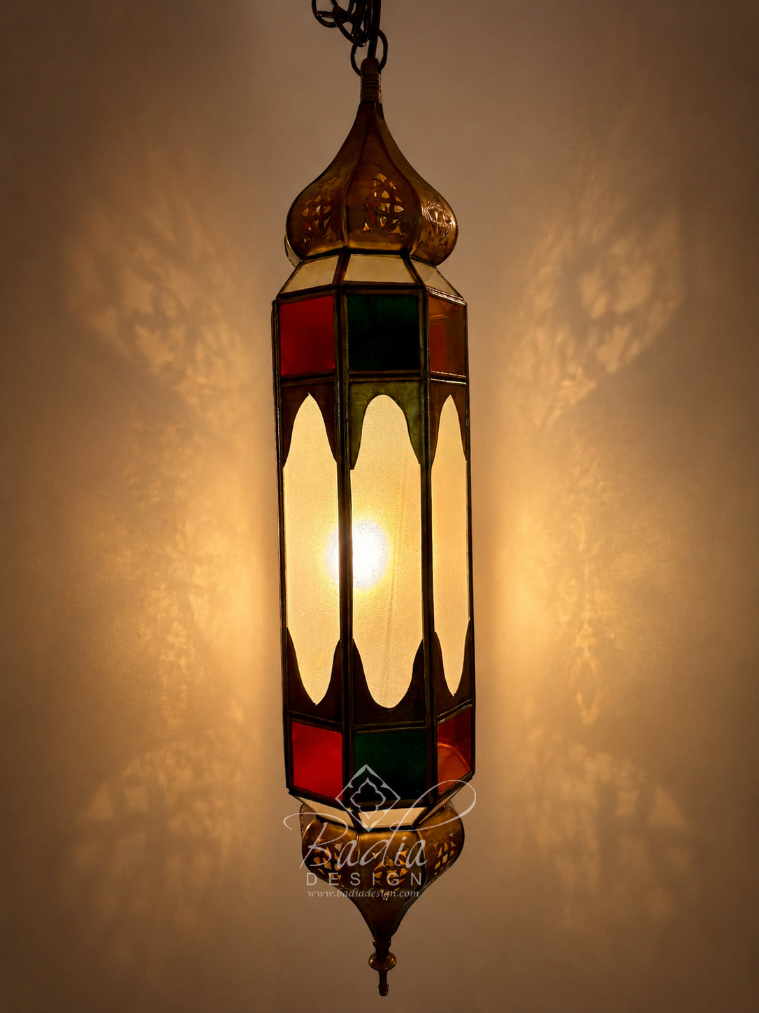 A hanging Moroccan-style stained glass lantern with a warm glowing light, featuring red, green, and cream glass panels framed by dark metal, shown against a warm beige wall with soft shadow projections.
