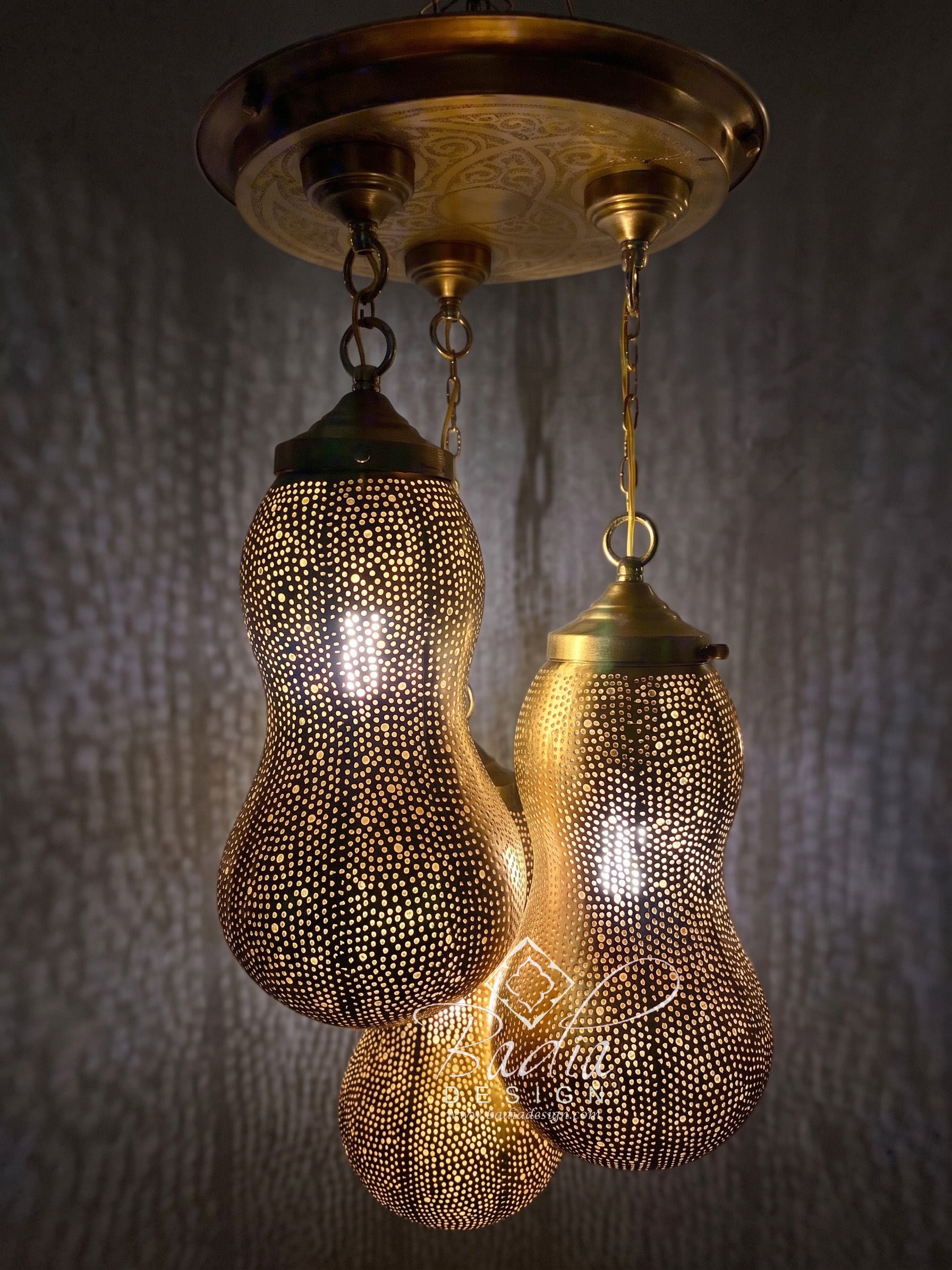 A hanging three-light ceiling fixture with brass-toned metal shades covered in small cutout dots, suspended from a round decorative ceiling canopy against a dark textured wall.