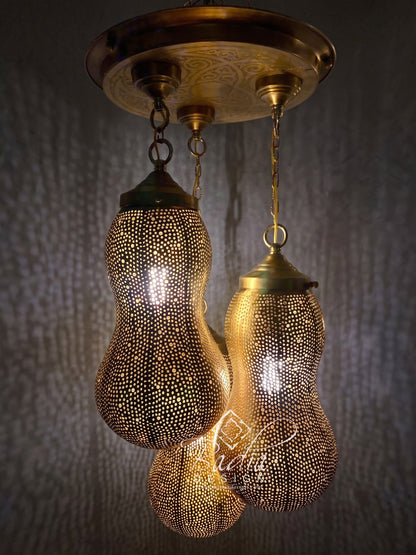 A hanging three-light ceiling fixture with brass-toned metal shades covered in small cutout dots, suspended from a round decorative ceiling canopy against a dark textured wall.